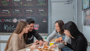 Un grupo de personas sentadas en una mesa, comiendo pizza. En la pared detrás hay una pizarra con varios escritos y dibujos.