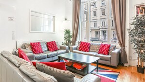 Una sala de estar moderna con sofás grises, cojines estampados en rojo y blanco, una mesa de centro y vistas a un paisaje urbano a través de grandes ventanales.