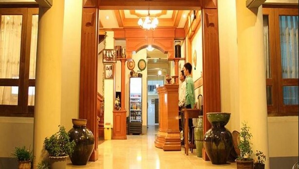 A hotel lobby with a wooden reception desk, a refrigerator, and potted plants. - Mr. Charles Hotel (Hsipaw)