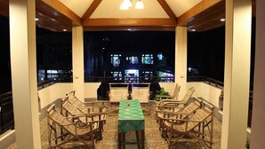 A covered outdoor seating area with wicker furniture and a table, overlooking a cityscape at night. - Mr. Charles Hotel (Hsipaw)