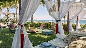 Un área de estar exterior lujosa con toldos blancos, sillones acolchados y vistas a la playa.