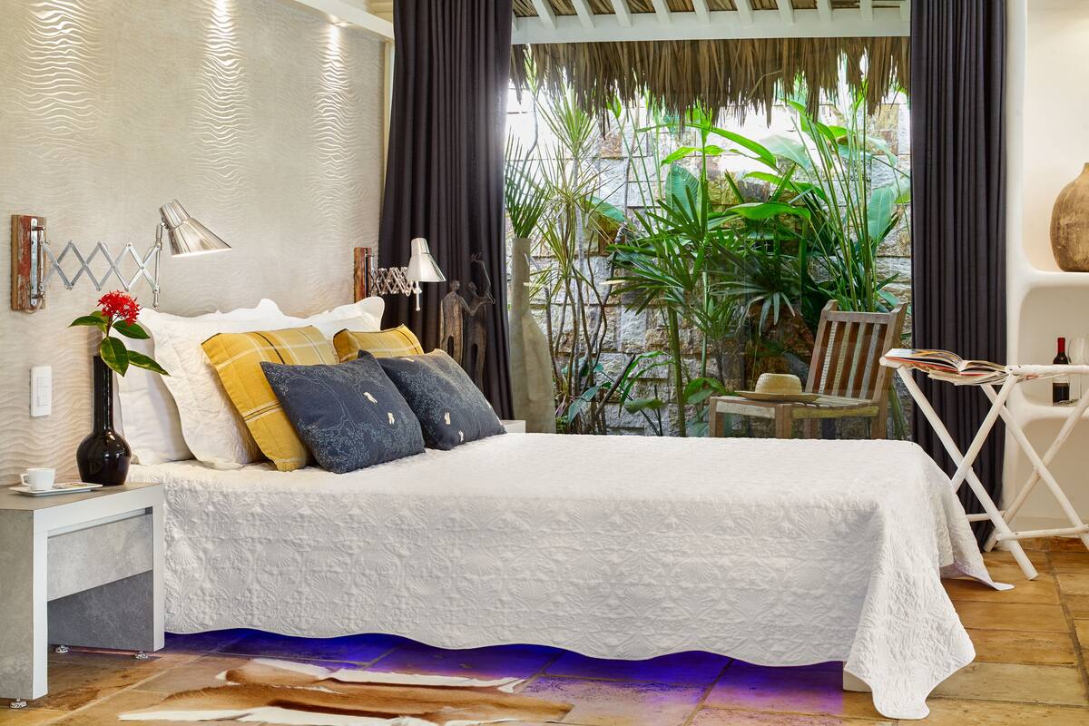 Una cama bien hecha con una colcha blanca, varios almohadones y una mesita de noche con una lámpara y un florero.