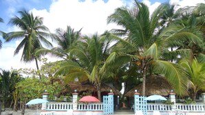 Una zona junto a la playa con estructuras de techo de paja, palmeras y sillas y mesas de plástico blanco.