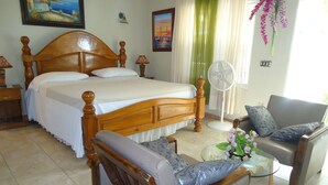 Un dormitorio con una cama de madera, una zona de estar con una silla y una mesita, y un ventilador.