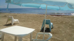 Una playa con una sombrilla grande, sillas de plástico blanco y una mesa.