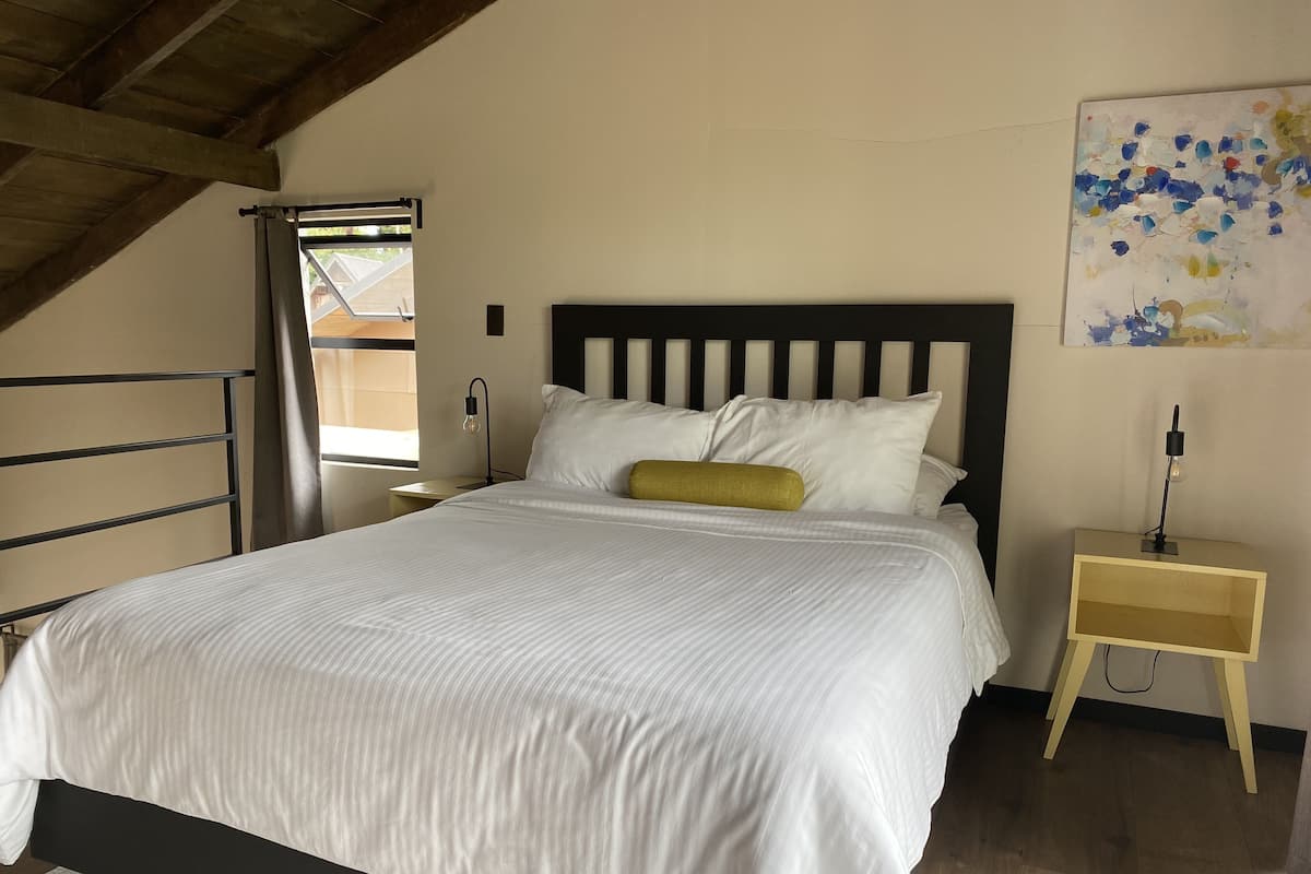A bedroom with a bed, a nightstand, a wall art piece, and a window with curtains.
