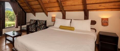 A bedroom with a wooden ceiling, a bed with white linens, two wall-mounted lamps, a small table with a plant, and a chair with patterned upholstery.