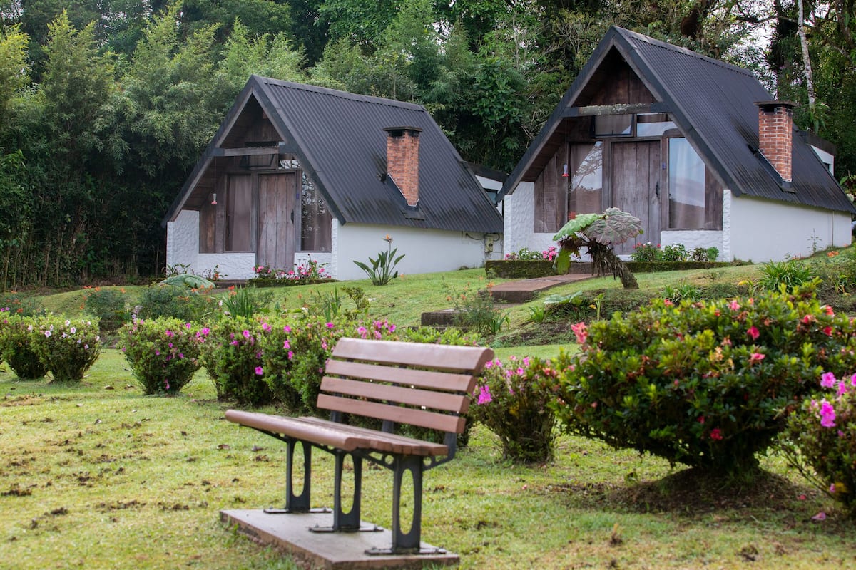 Two rustic cottages with sloped roofs and brick chimneys, surrounded by well-maintained gardens and a wooden bench in the foreground.