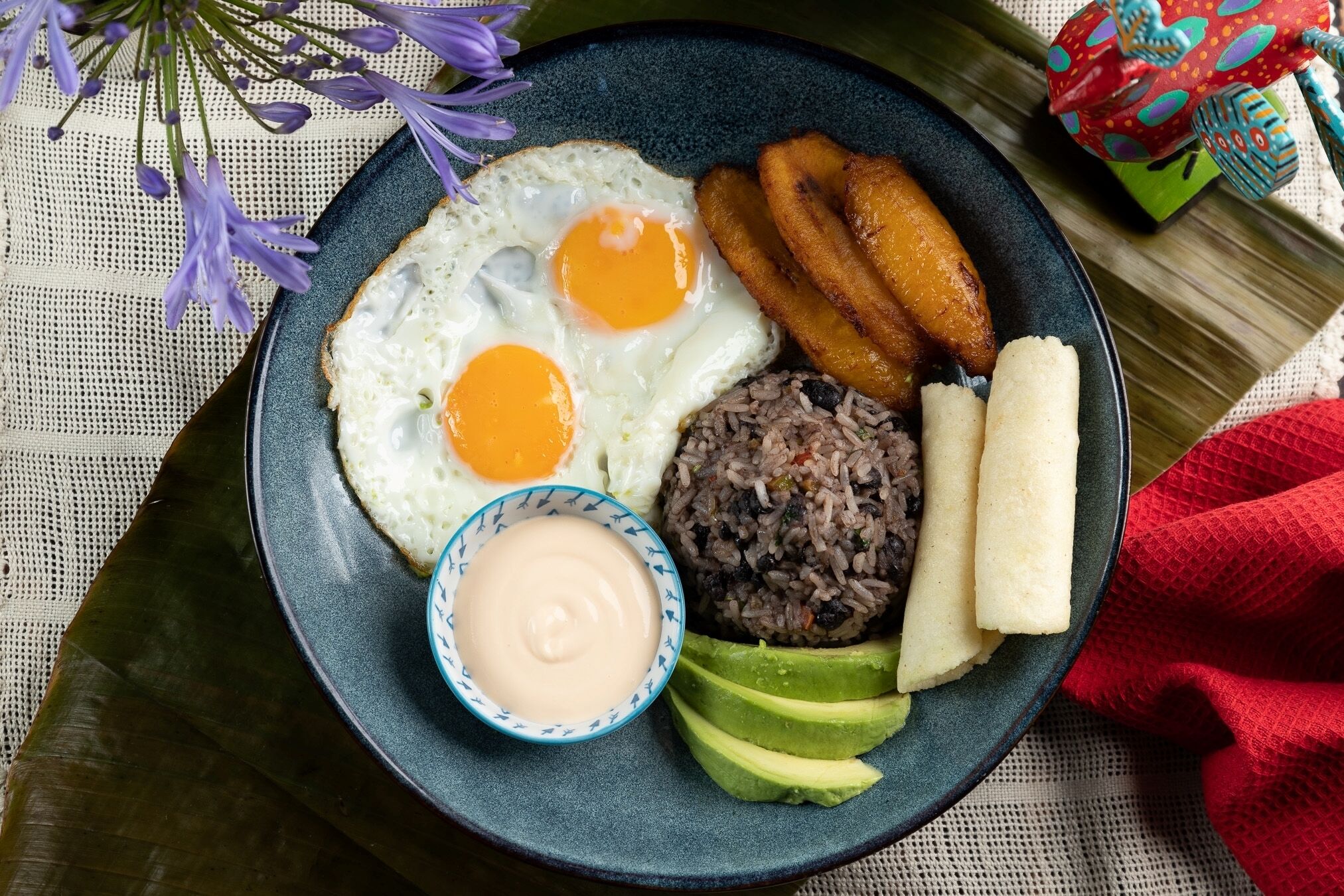 A plate with fried eggs, black rice, plantains, avocado, and a small bowl of sauce.