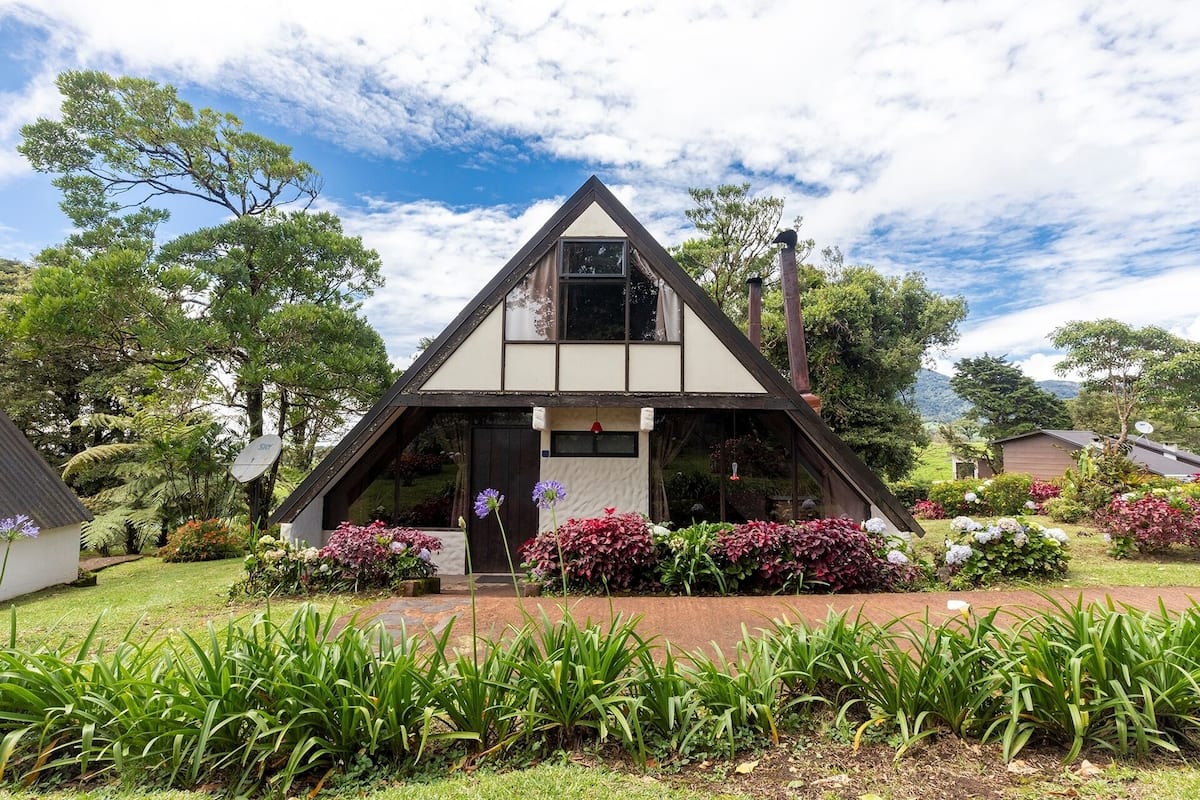 A triangular-roofed building with a visible front porch and a garden with various plants and flowers.