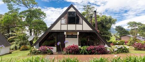 A triangular-roofed building with a visible front porch and a garden with various plants and flowers.