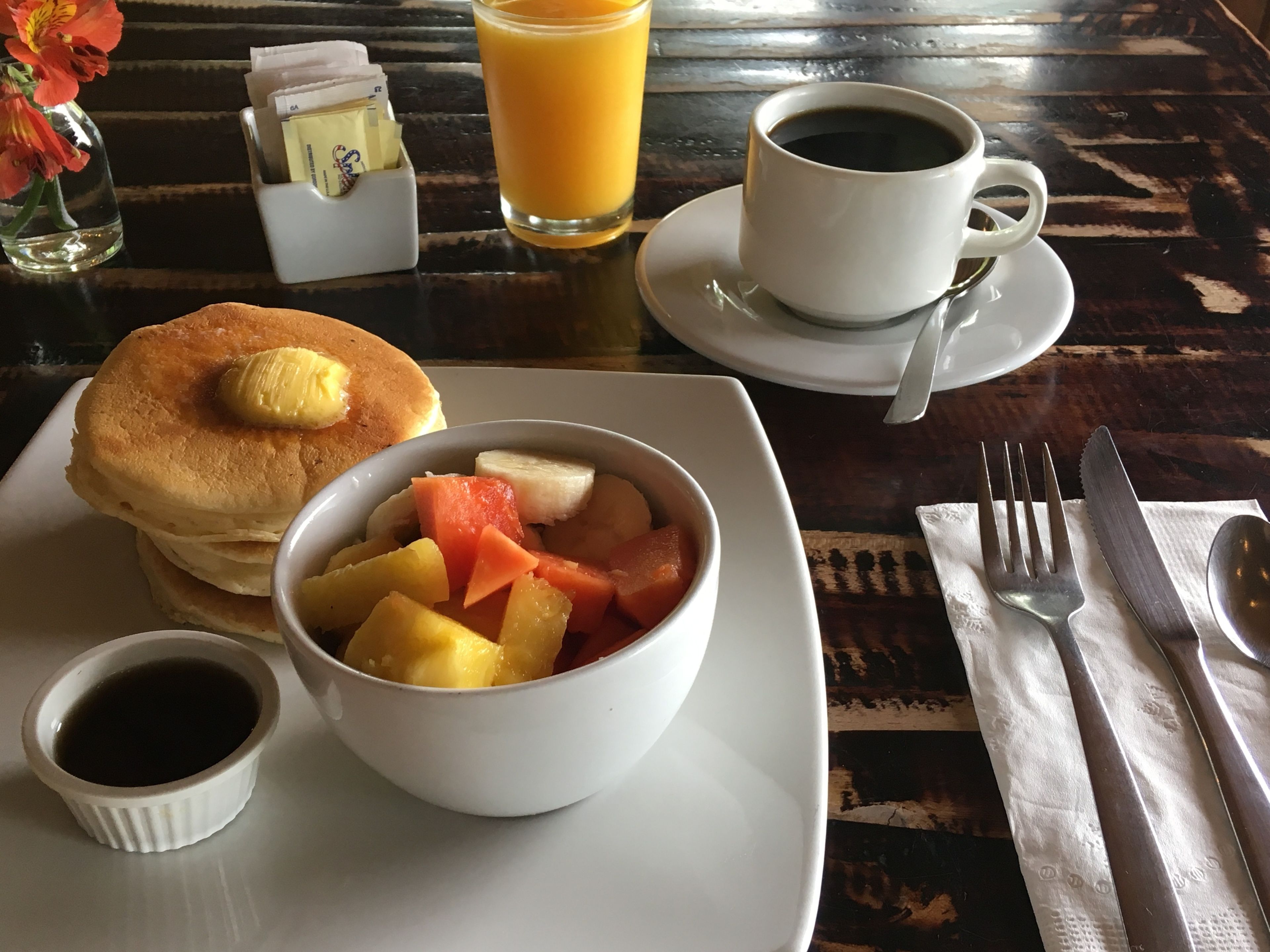 A breakfast table with pancakes, a fruit bowl, a cup of coffee, and a glass of orange juice.