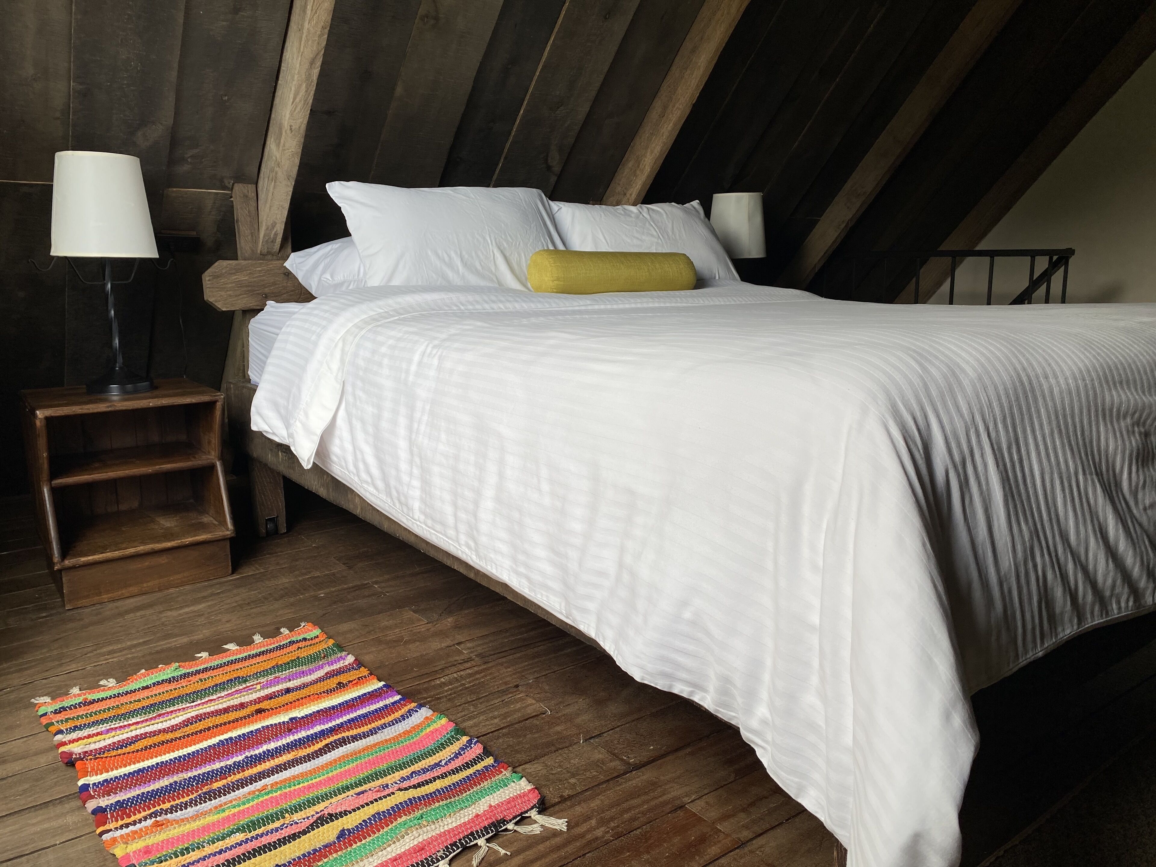 Een slaapkamer met een houten plafond, een bed met wit beddengoed, een kleurrijk vloerkleed en twee bedlampjes.