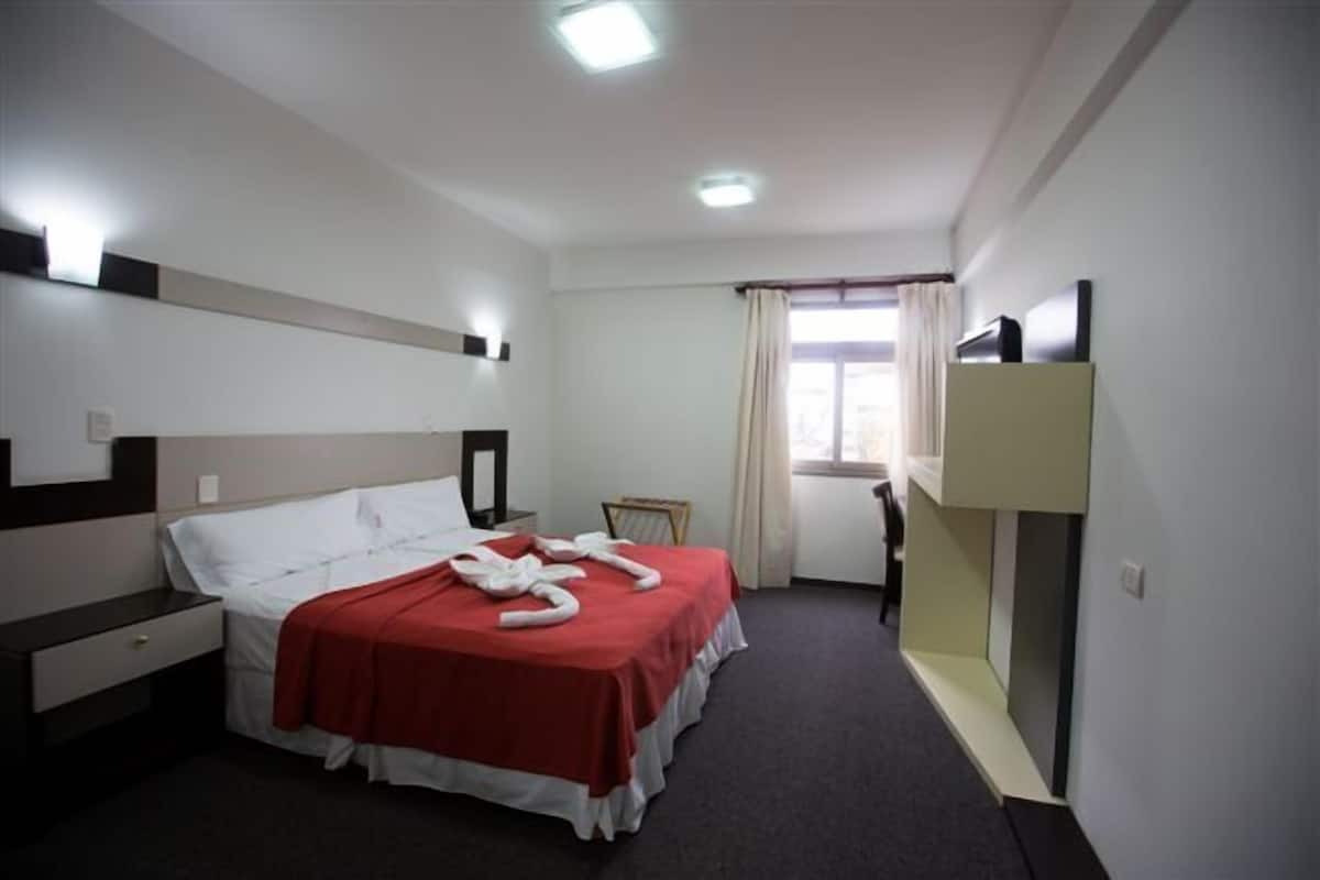 A hotel room with a bed, a desk, a chair, a window with curtains, and a built-in shelf.