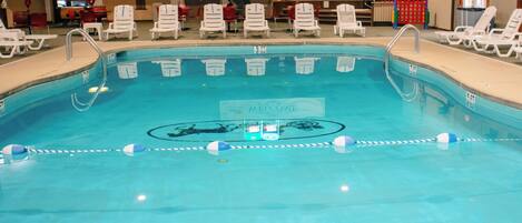An indoor swimming pool with clear lane dividers, a welcome sign, and white lounge chairs around the perimeter.