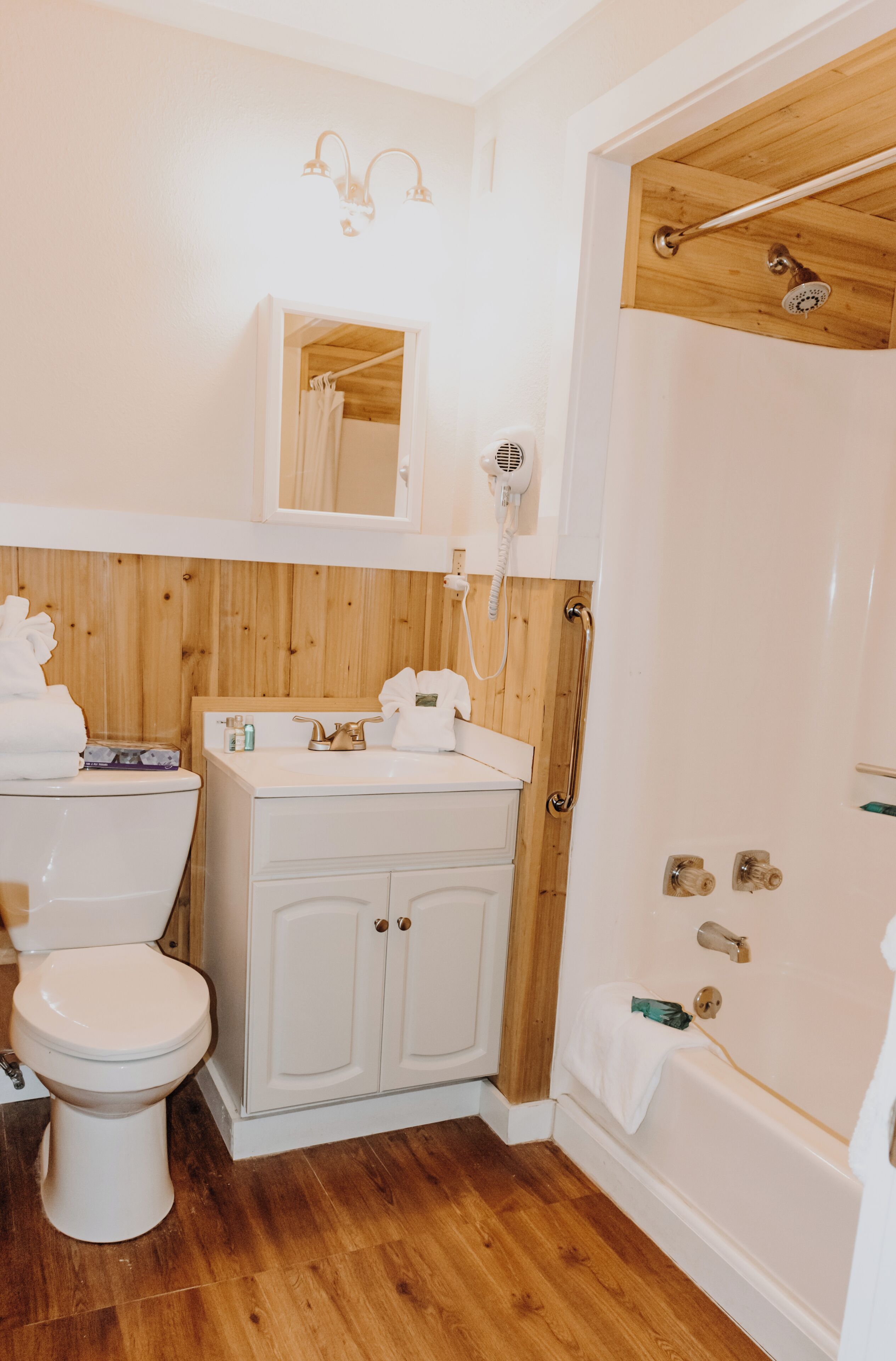 A bathroom with a wooden floor, a white sink, a toilet, a shower, and a mirror above the sink.