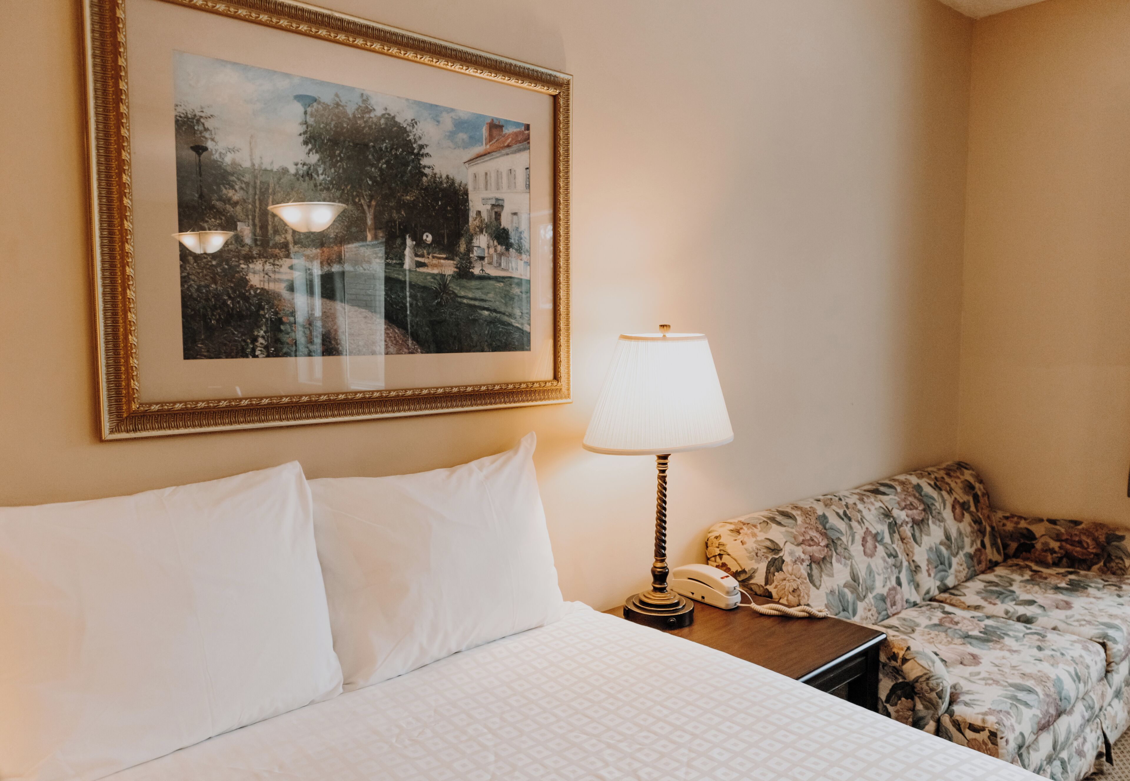 A hotel room with a bed, a framed landscape painting, a lamp, and a floral sofa.