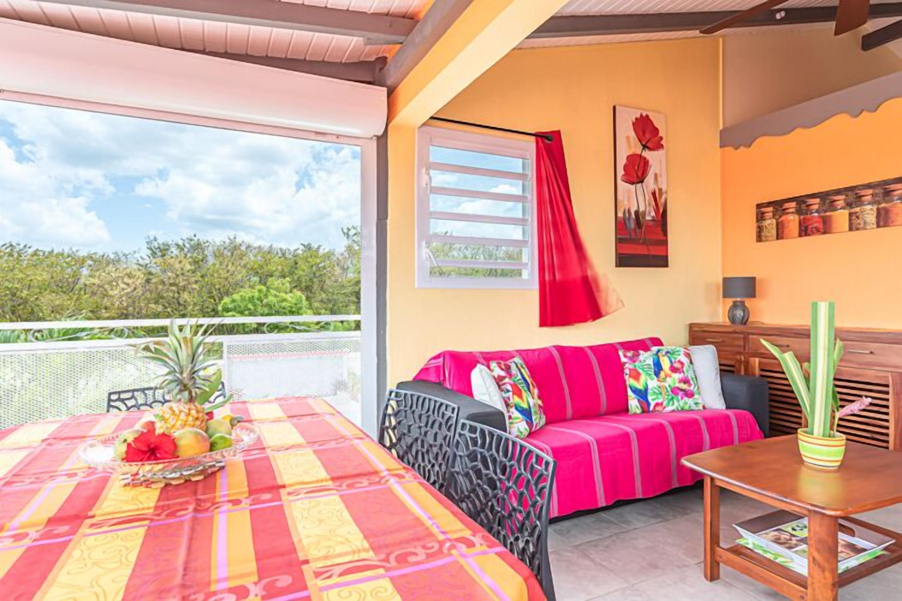 A room with a balcony, a table with fruit and a bottle, a sofa with colorful cushions, and a wooden cabinet.