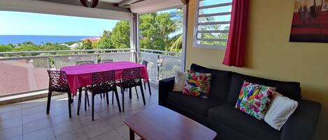 Un balcón con mesa y sillas, un sofá con cojines estampados y vistas al mar.