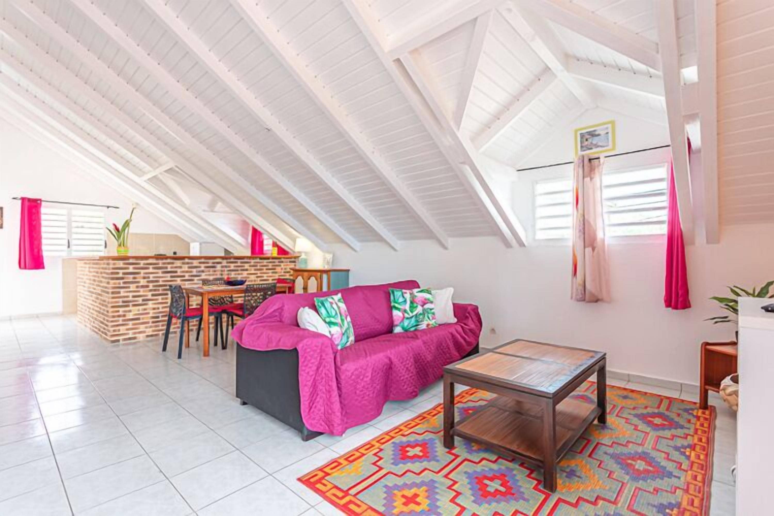 A spacious loft with a high ceiling, a cozy seating area with a sofa and a coffee table, a kitchen area with a brick wall, and a dining table with chairs.