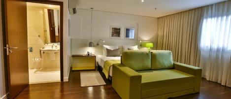 Habitación de hotel con suelo de madera, sofá verde, cama con ropa de cama blanca, mesita de noche, lámpara, espejo y baño con bañera y lavabo.