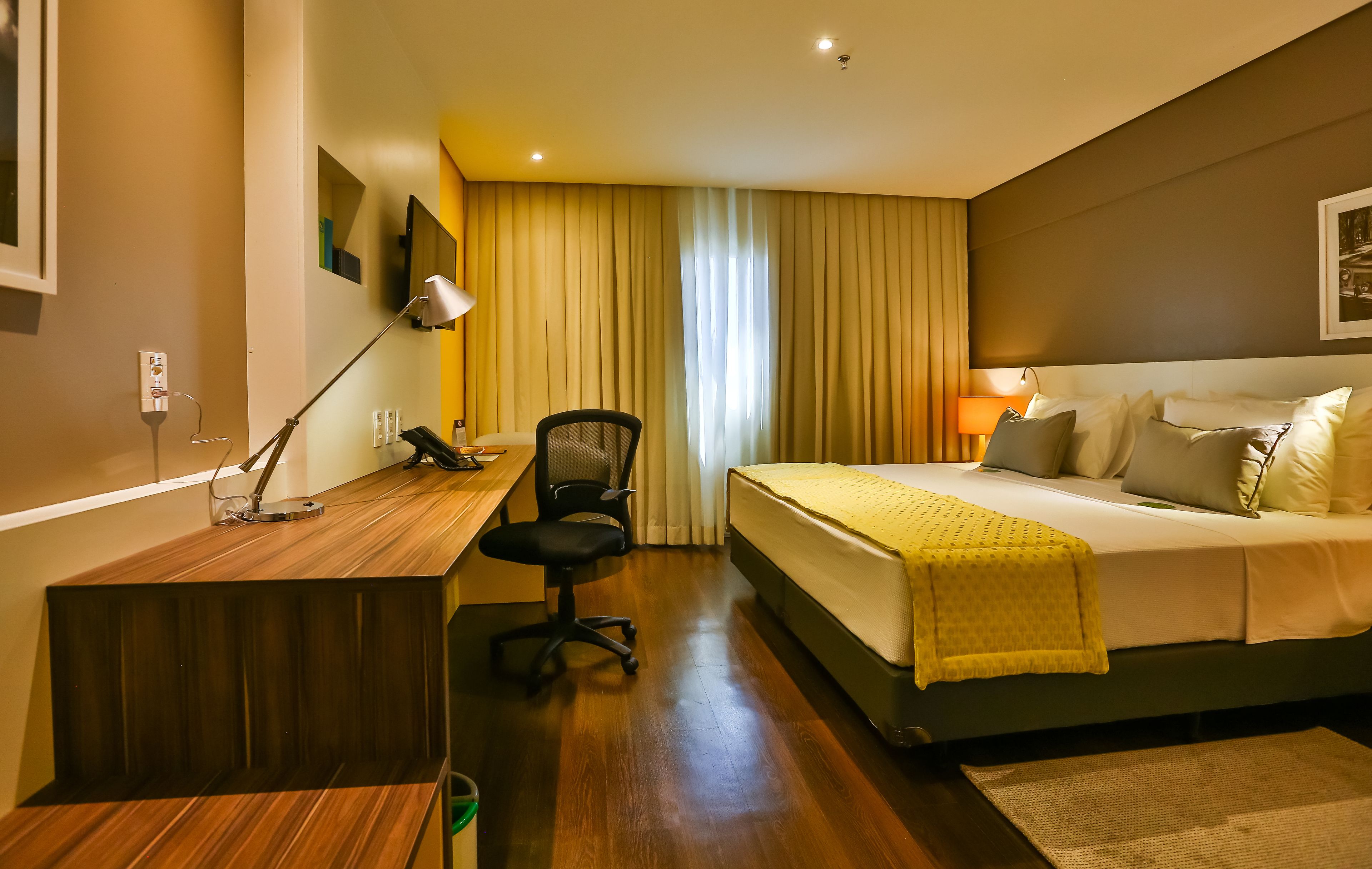 Habitación de hotel con un escritorio de madera, una silla, una cama con manta amarilla y un ventanal con cortinas.