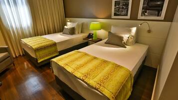 Quarto de hotel com duas camas, uma mesa de cabeceira com abajur e uma janela com cortinas.