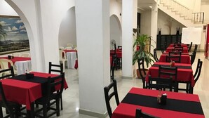 Un restaurant avec des tables recouvertes de nappes rouges et des chaises noires, des piliers blancs et un escalier en arrière-plan.