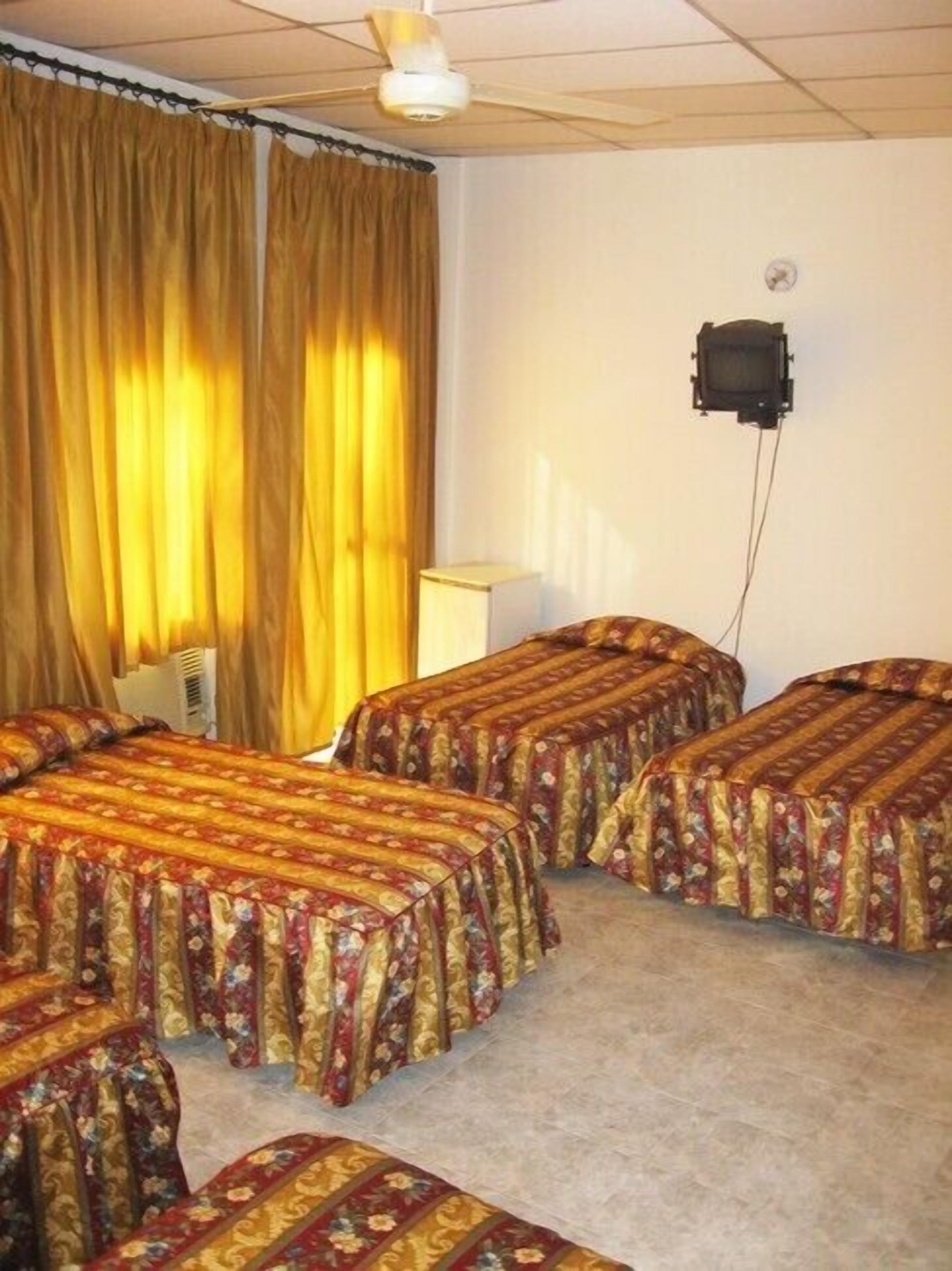 A room with four beds, each with floral-patterned bedding, a ceiling fan, and a television mounted on the wall.