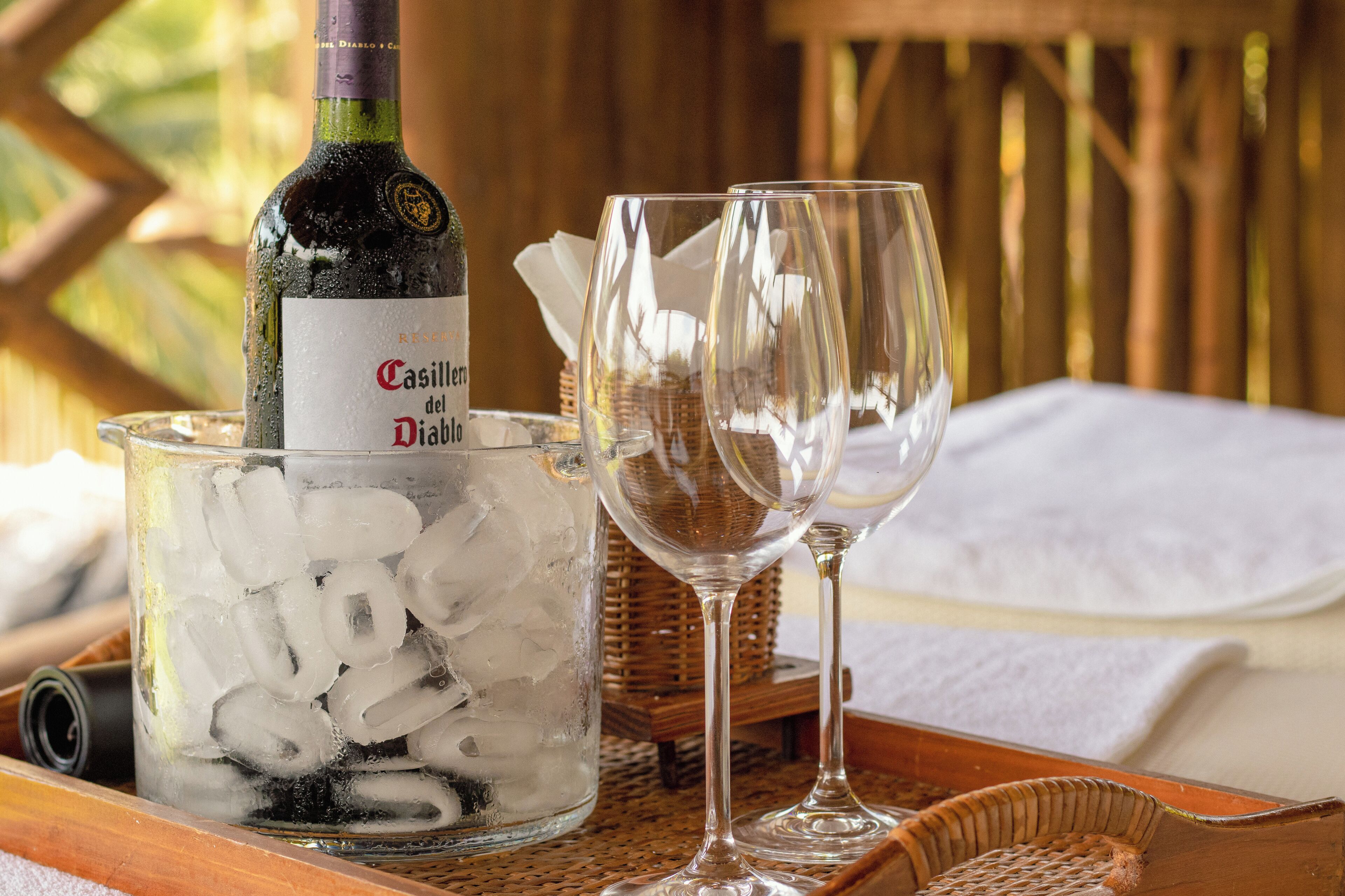A bottle of Casillero del Diablo wine in a glass container with ice, two empty wine glasses, and a wicker basket on a wooden tray.