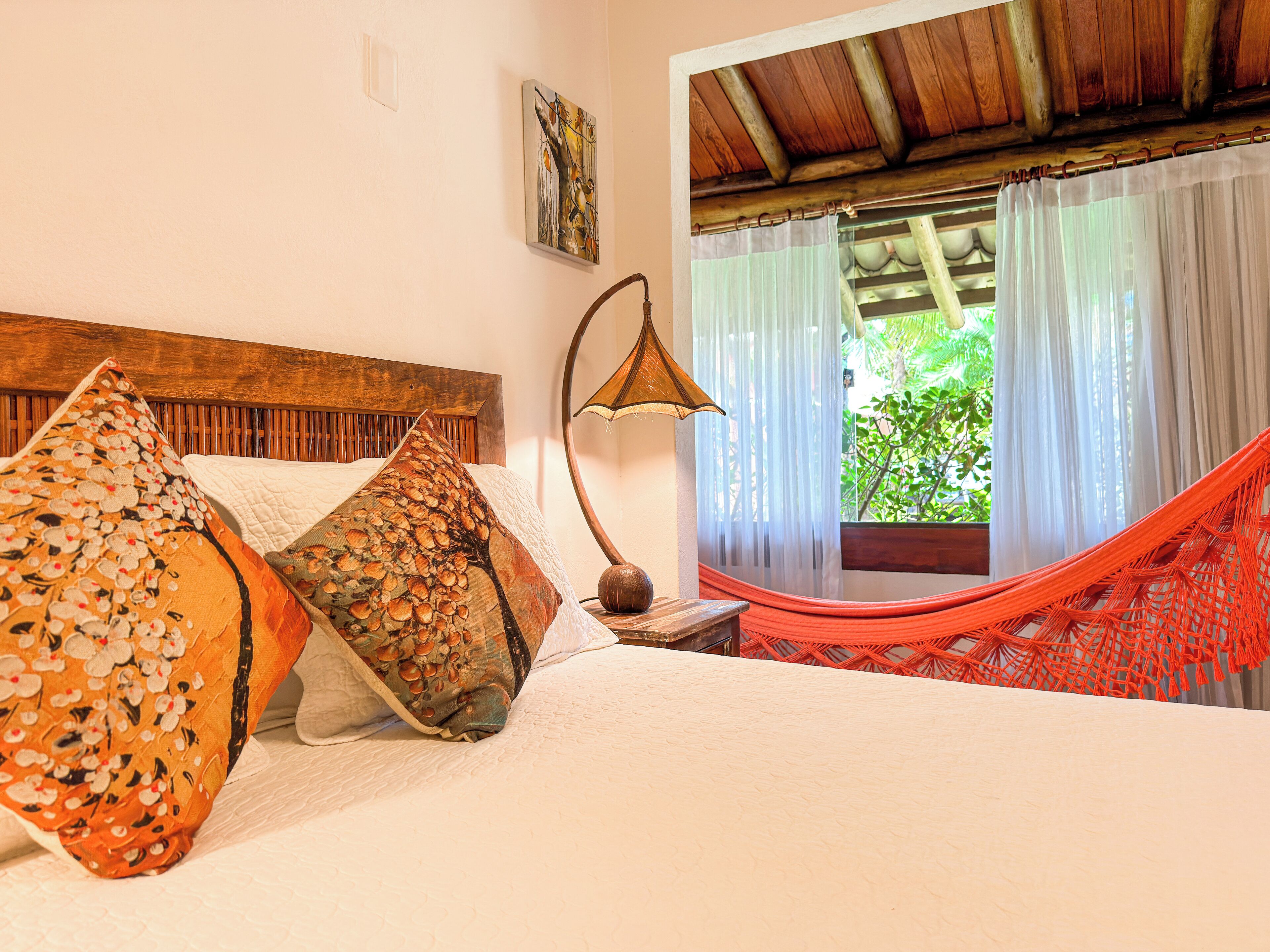 A bed with floral pillows, a wooden headboard, a bedside lamp, and a view of a hammock and greenery through a window.