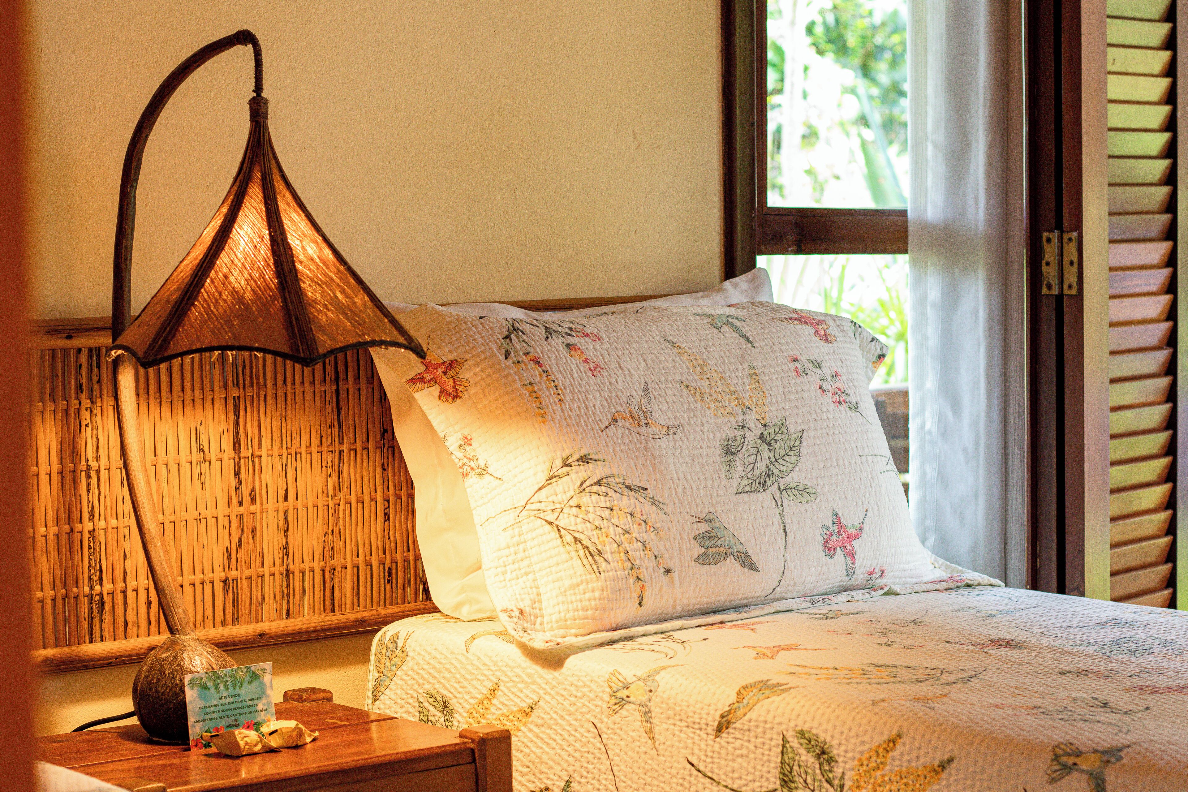 A bed with floral bedding, a hanging lamp, and a wooden nightstand with a vase and a card.