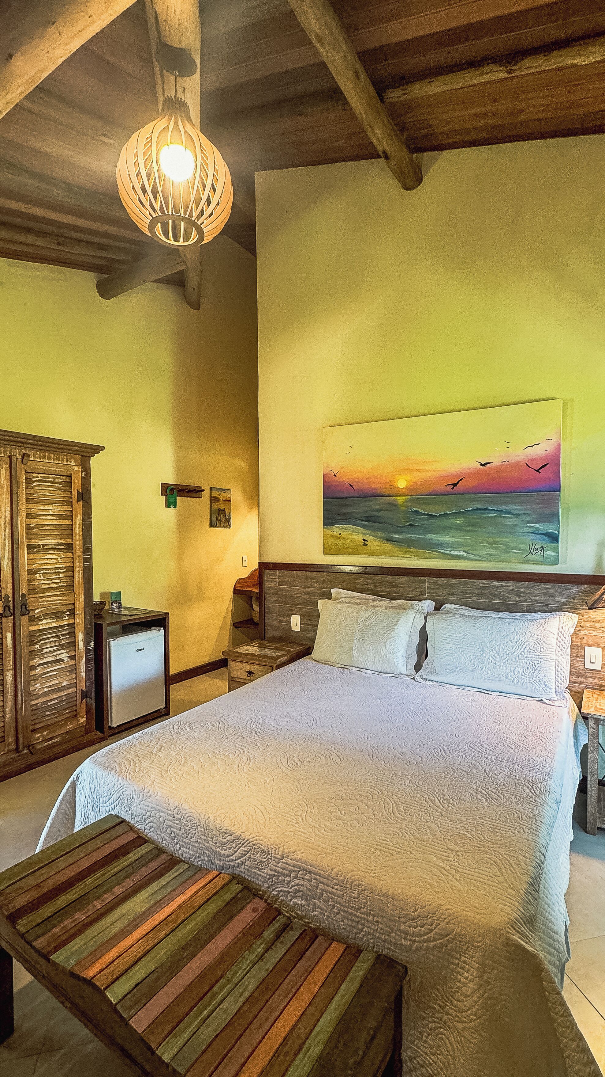 Ein Schlafzimmer mit einem Bett, einer hölzernen Bank, einem Sonnenuntergangsbild und einer Deckenleuchte.