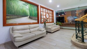 A modern hotel lobby with a beige leather sofa, a large bamboo forest painting, a glass partition with a geometric design, and a staircase with a metal railing.