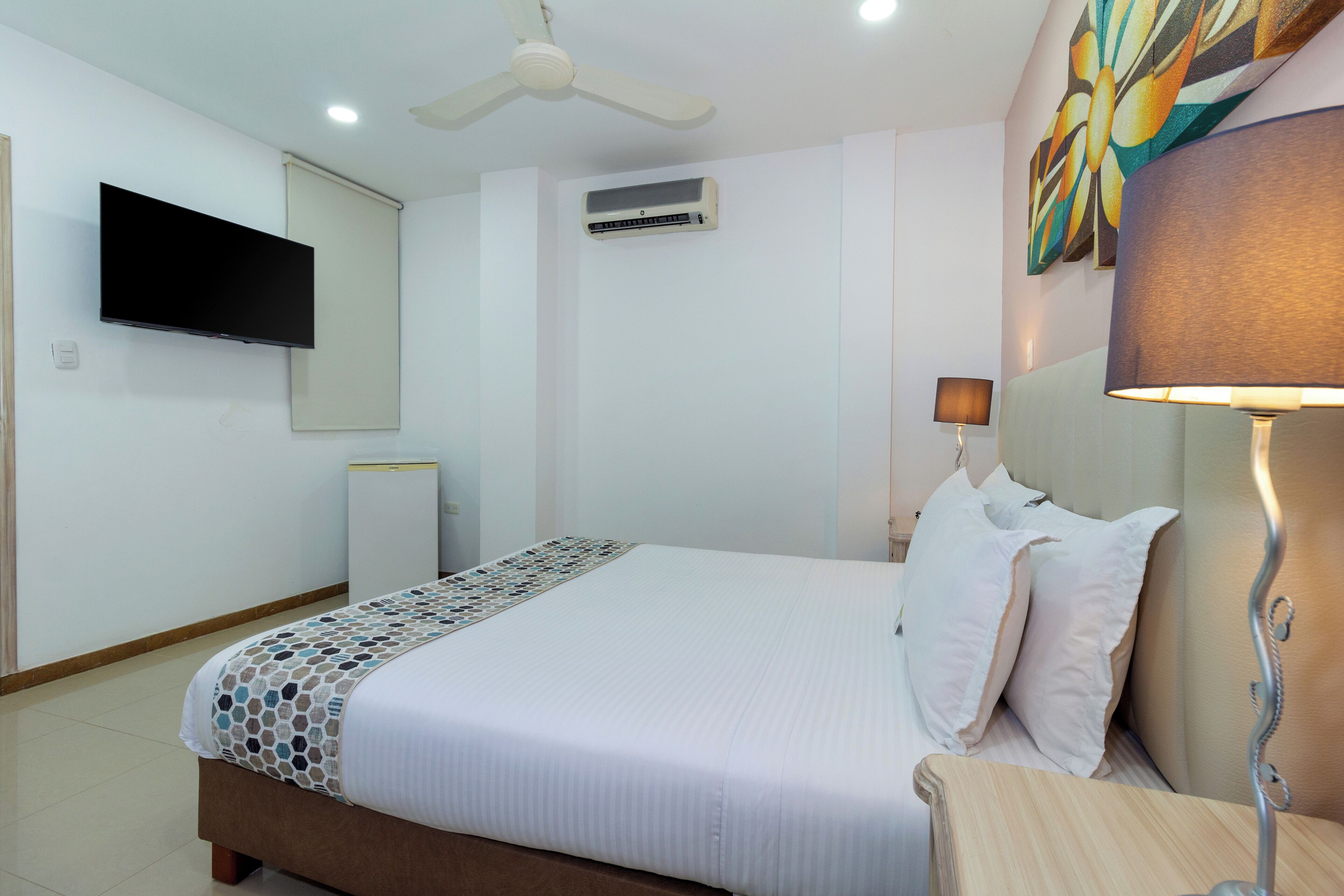 Una habitación de hotel con una cama, un televisor montado en la pared, un equipo de aire acondicionado, un ventilador de techo, una lámpara de noche y un cuadro en la pared.