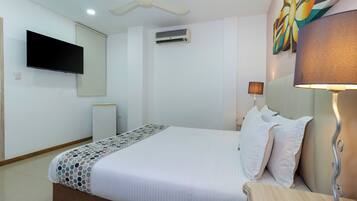 Una habitación de hotel con una cama, un televisor montado en la pared, un equipo de aire acondicionado, un ventilador de techo, una lámpara de noche y un cuadro en la pared.