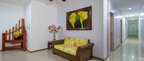 A hallway with a wicker sofa, a wooden chair, a ceiling fan, a framed painting, and a vase with flowers.