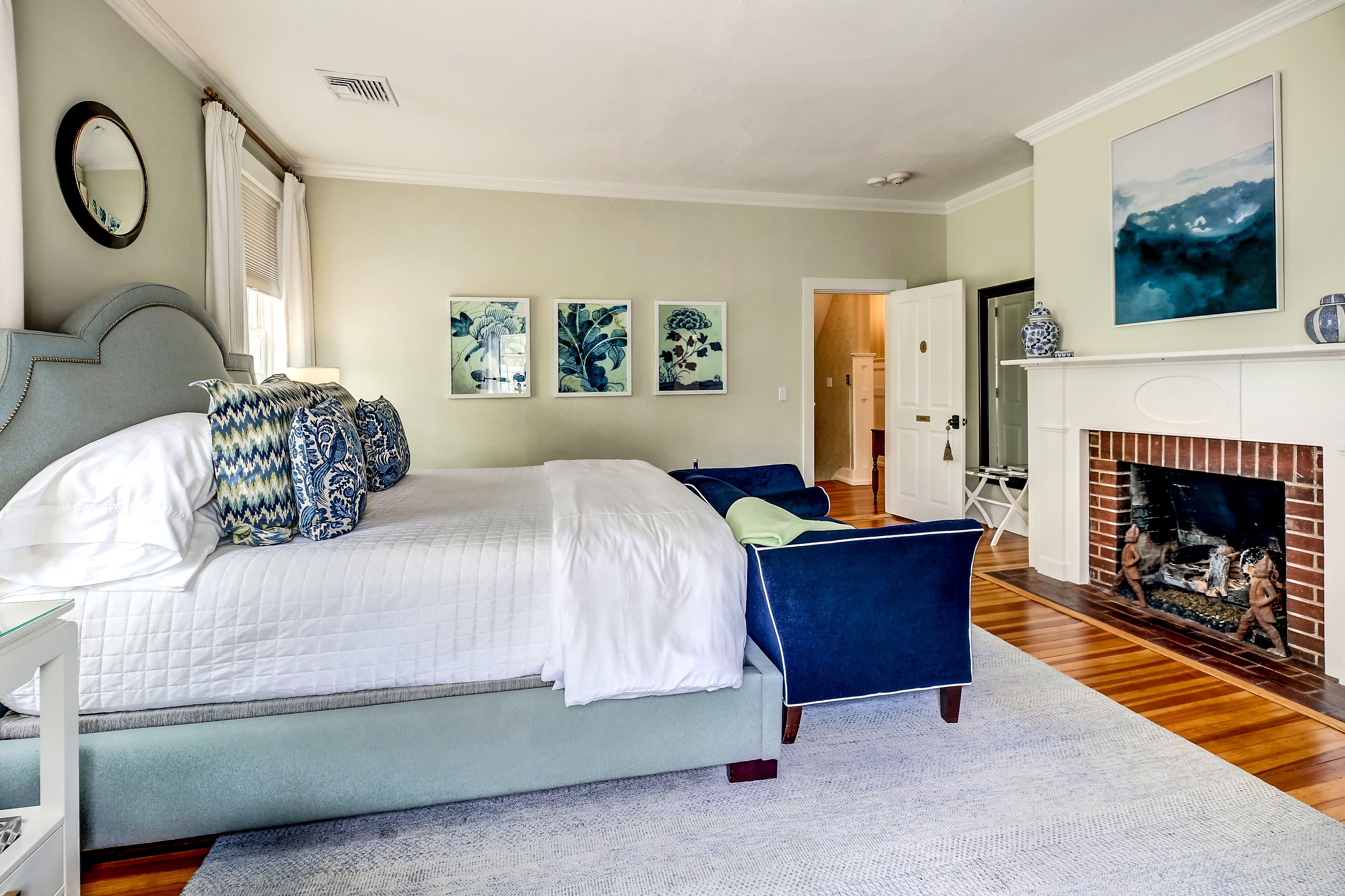 A bedroom with a large bed, a blue chair, a fireplace, and three framed artworks on the wall.