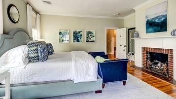 A bedroom with a large bed, a blue chair, a fireplace, and three framed artworks on the wall.