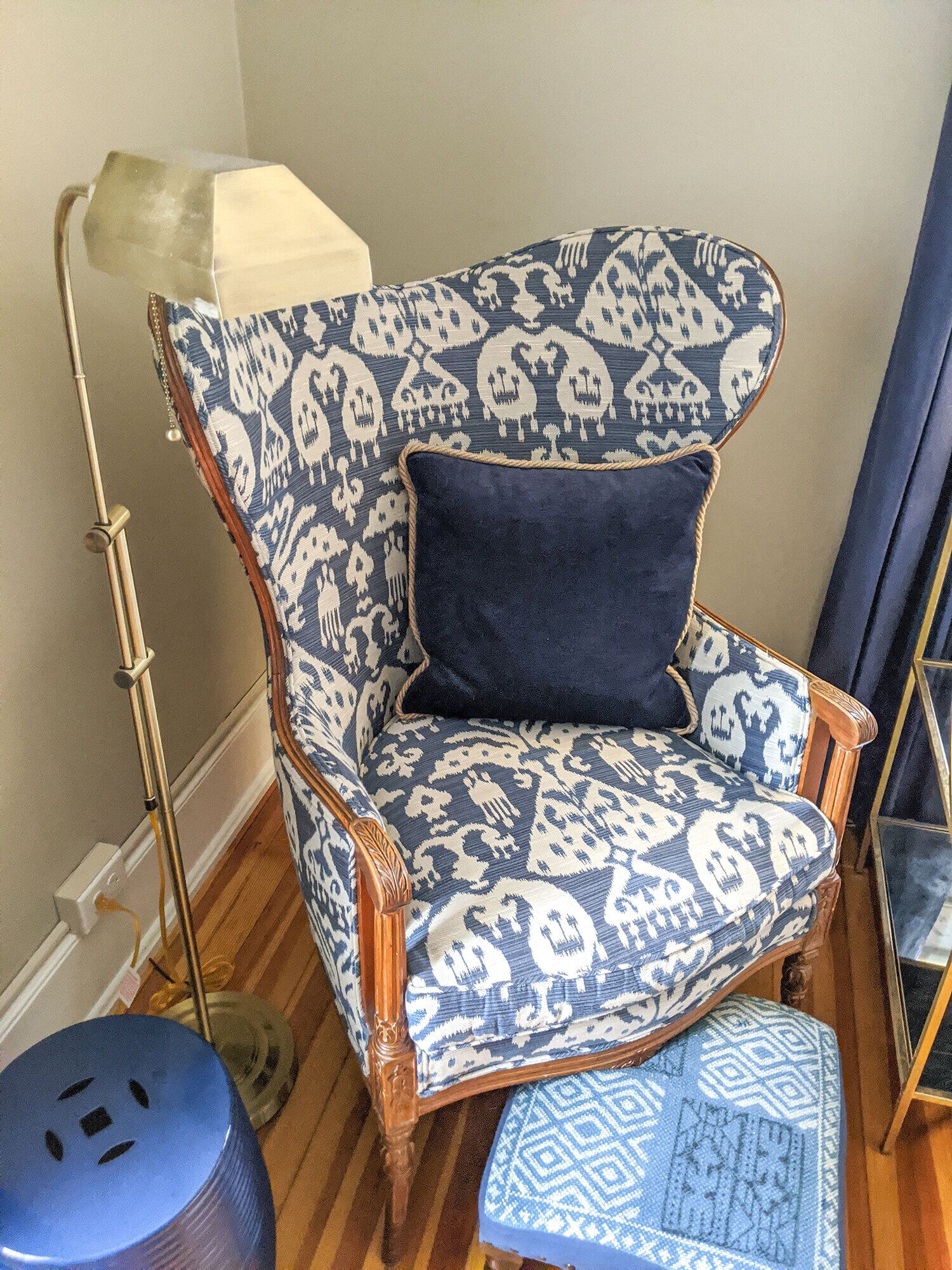 A chair with a patterned fabric and a dark cushion, placed in a room with wooden flooring and a lamp.