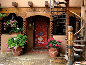 Entrada de un edificio con una escalera de madera, una puerta roja con motivos florales y plantas en macetas con flores rosas.