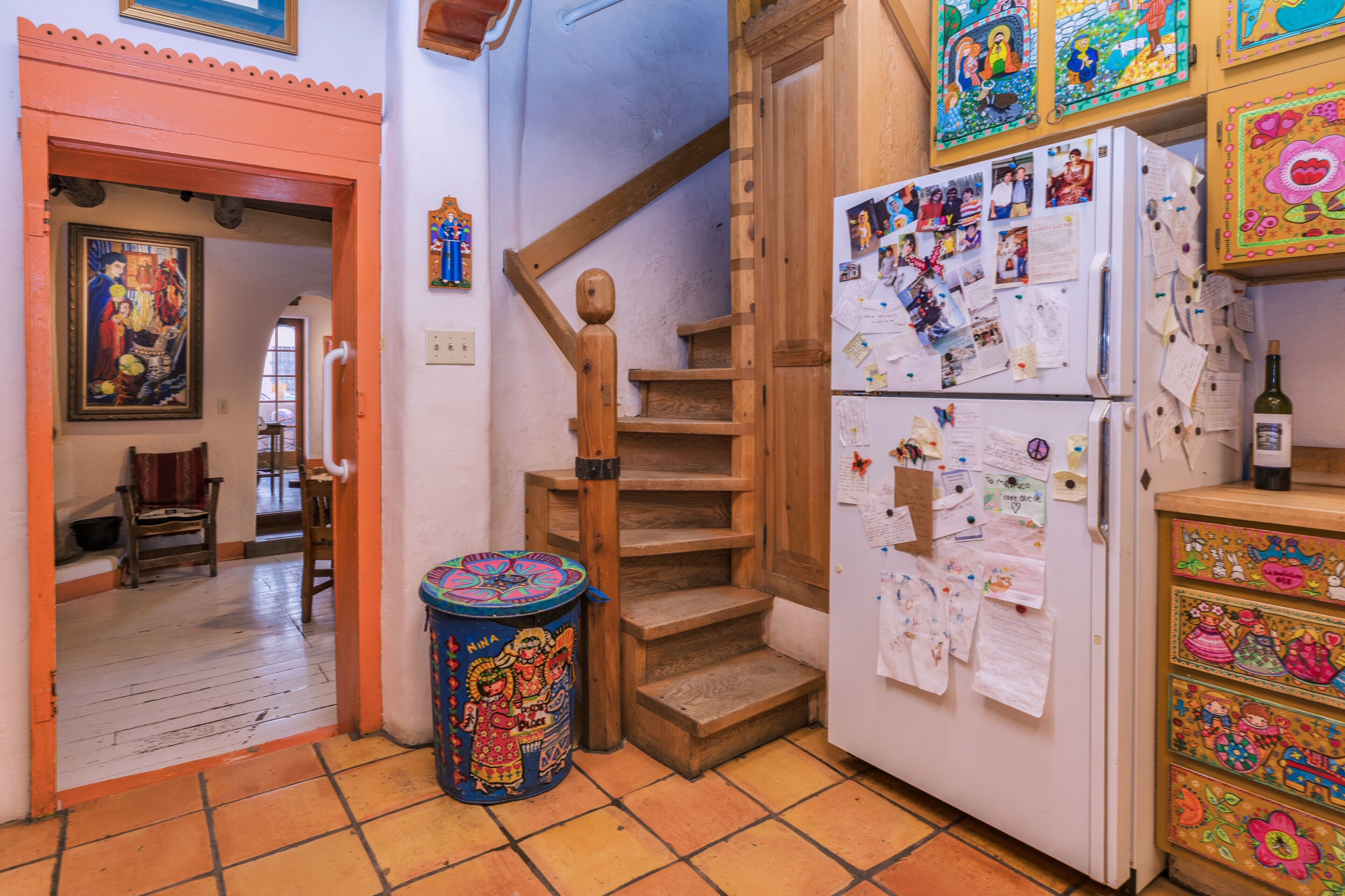 Una cocina con una escalera de madera, un refrigerador lleno de notas e imágenes, y un cubo de basura colorido.