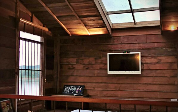 Une cabane en bois avec un téléviseur fixé au mur, une fenêtre donnant sur l’extérieur et une petite table sur laquelle est posée une photo encadrée.