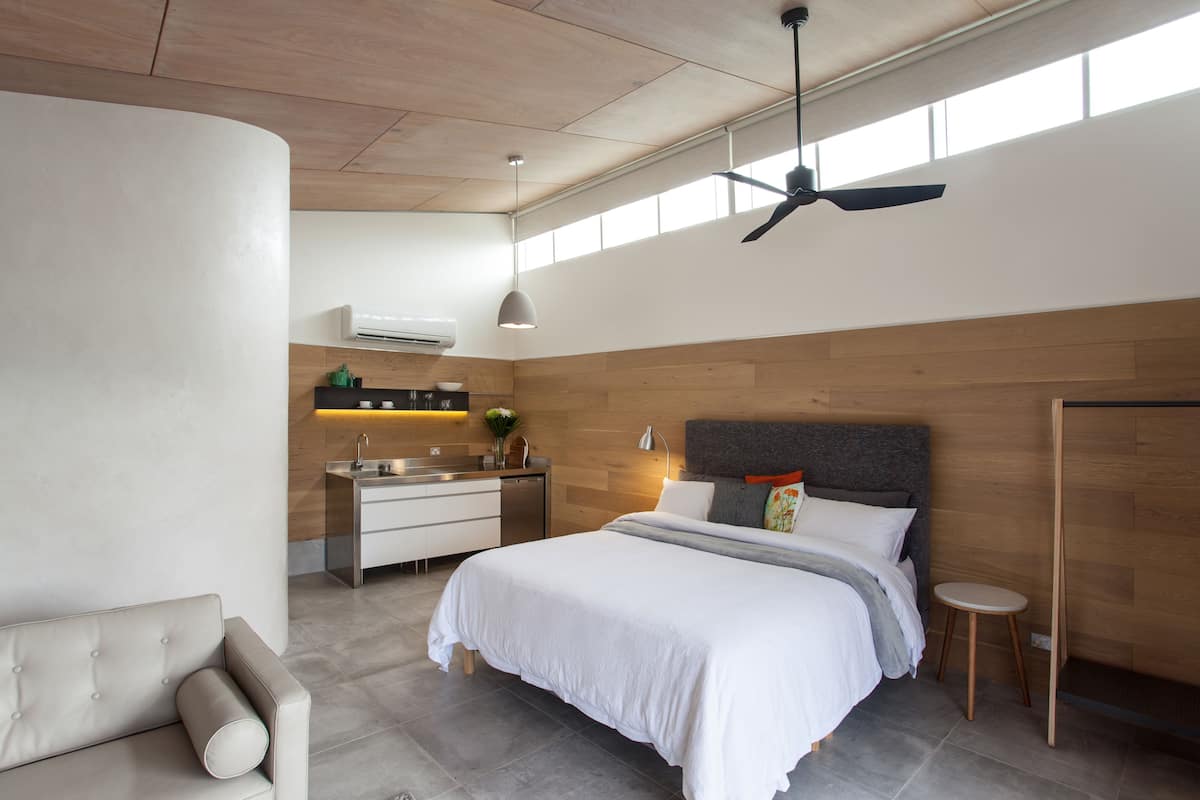 Un dormitorio moderno con una cama grande, una mesita de noche pequeña, un escritorio empotrado con una silla, un ventilador de techo y una ventana que proporciona luz natural.