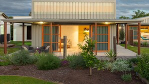 Ein modernes Haus mit großer Glasschaufensterfront, einer überdachten Terrasse und einem Garten mit verschiedenen Pflanzen.