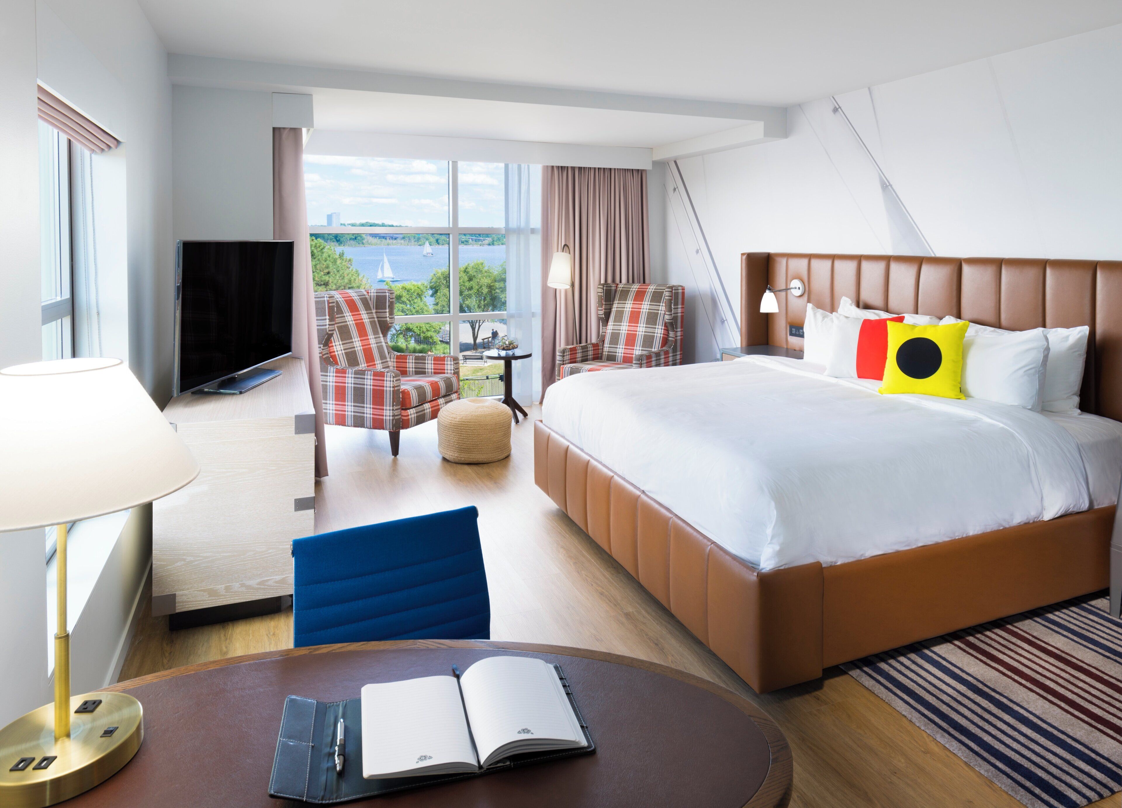 Habitación de hotel con cama, dos sillones, televisor, escritorio con lámpara y vistas al exterior.