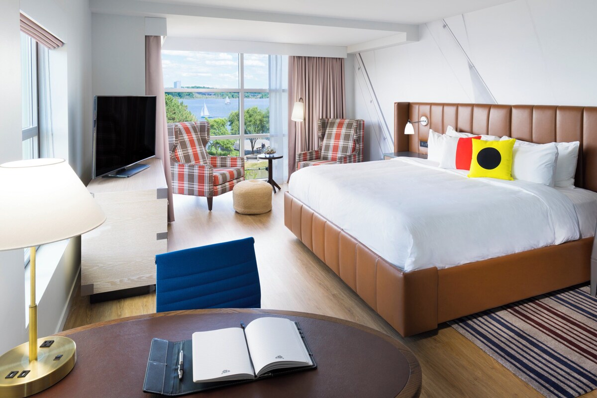 Une chambre d’hôtel comprenant un lit, deux fauteuils, une télévision, un bureau avec une lampe et une vue sur l’extérieur.