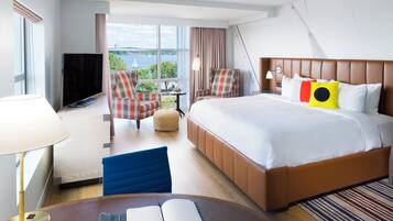 A hotel room with a bed, two armchairs, a TV, a desk with a lamp, and a view of the outdoors.