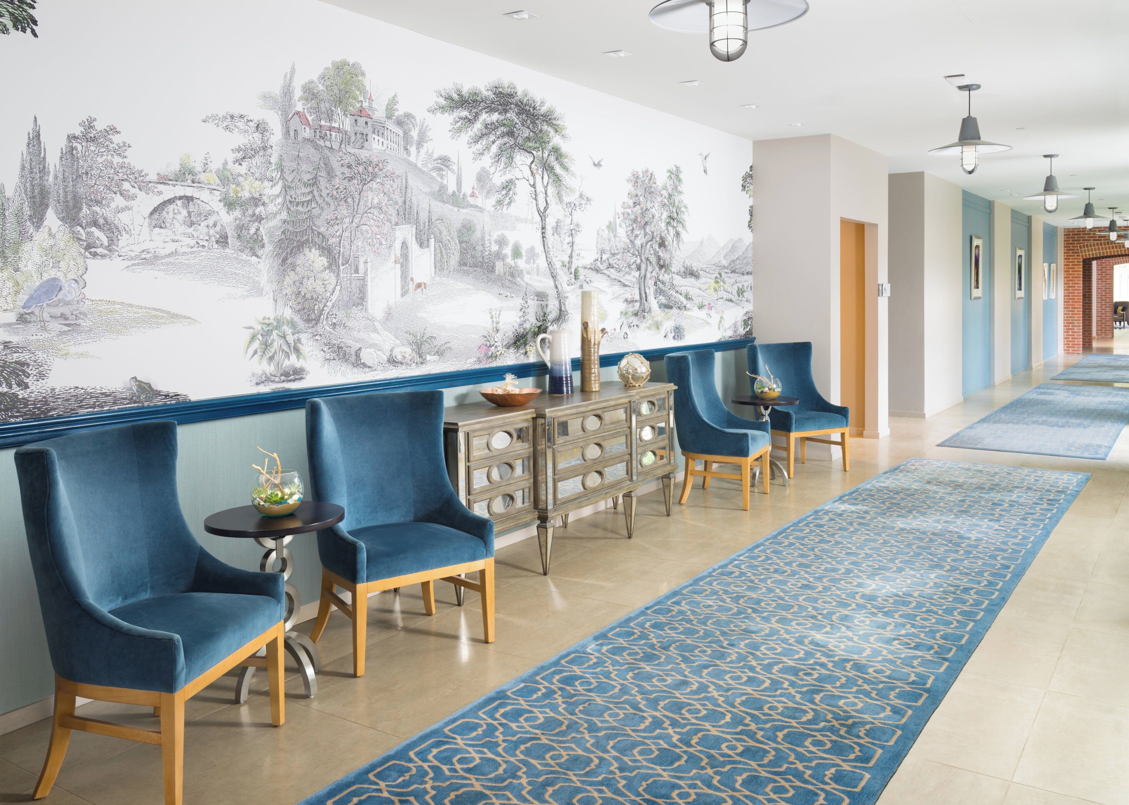 Eine Hotelhalle mit einer großen Wandmalerei, blauen Polsterstühlen, einem Holzschrank und einem gemusterten Teppich.