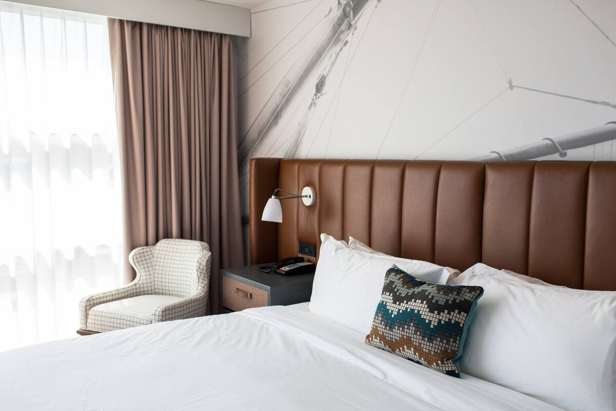 Ett hotellrum med en stor säng, ett nattduksbord med en lampa, en stol och utsikt över omgivningarna.